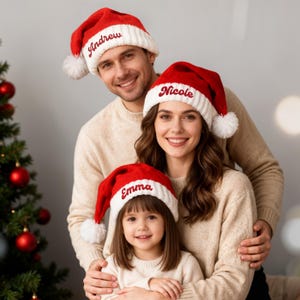 Könnte beinhalten: Eine Familie trägt passende rot-weiße Weihnachtsmützen mit gestickten Namen. Die Mützen haben einen weißen Bommel und werden von einem Mann, einer Frau und einem kleinen Mädchen getragen. Sie lächeln vor einem Weihnachtsbaum.