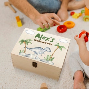 Könnte beinhalten: Eine hölzerne Schatzkiste mit dem Text "Alex's Treasure Box" in Grün. Die Kiste zeigt eine Aquarellillustration eines Dinosauriers, Palmen und Wolken. Die Kiste ist rechteckig mit einem Metallverschluss.