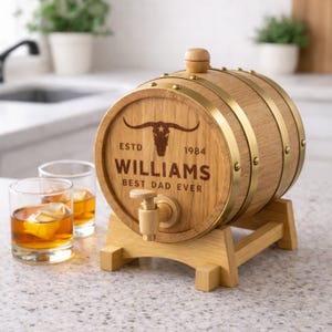 May include: A wooden barrel with brass bands and a wooden stand. The barrel has the text "ESTD 1984 WILLIAMS BEST DAD EVER" and a longhorn skull graphic. Two glasses of amber liquid with ice cubes are in the foreground.