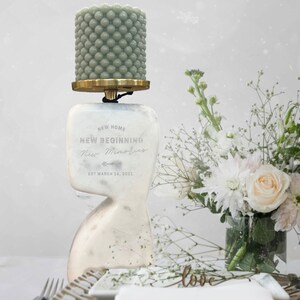May include: White marble candle holder with a green beaded candle on top. The holder has the text "NEW HOME NEW BEGINNING New Memories EST MARCH 16, 2021" engraved on it.