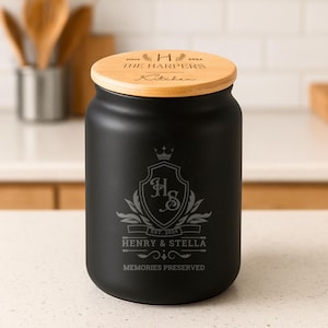 Op de afbeelding: Een zwarte keukencollectiepot met een houten deksel. Het deksel is gegraveerd met "since 2024 THE HARPERS Kitchen". De pot heeft gegraveerde tekst: "HENRY & STELLA EST. 2024 MEMORIES PRESERVED" en een wapen met de initialen "H & S".