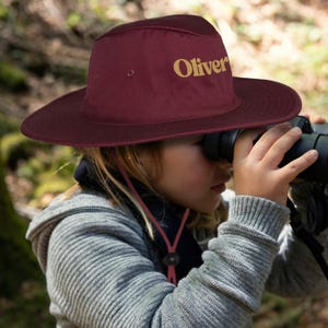 May include: A burgundy sun hat with the name "Oliver" in gold lettering. The hat has a wide brim and a chin strap. The person is wearing a grey hooded sweatshirt and looking through binoculars.