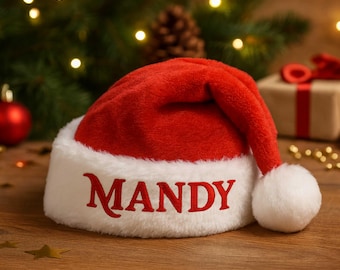 May include: A red Santa hat with a white trim and a white pom-pom. The name "MANDY" is embroidered in red on the white band. The hat is on a wooden surface with Christmas decorations in the background.
