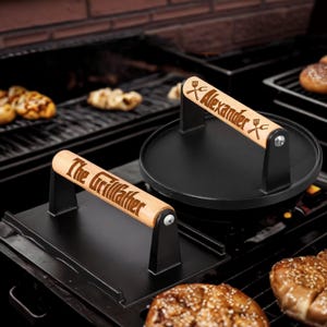 May include: Two black cast iron grill presses with wooden handles. One is rectangular with "The Grillfather" engraved. The other is round with "Alexander" engraved. Both are on a grill with cooked food, ideal for grilling burgers.