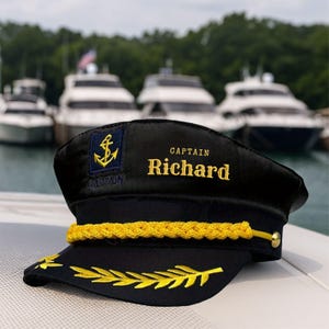 May include: Black captain's hat with gold embroidered details. The hat features an anchor emblem, the name "CAPTAIN" and the name "Richard" in gold lettering. A gold rope trim and gold leaf embroidery adorn the hat's brim. Boats are visible in the background.