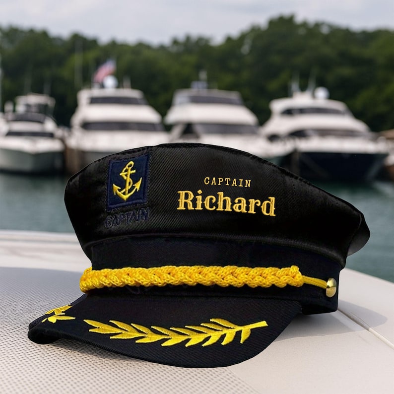 May include: Black captain's hat with gold embroidered details. The hat features an anchor emblem, the name "CAPTAIN" and the name "Richard" in gold lettering. A gold rope trim and gold leaf embroidery adorn the hat's brim. Boats are visible in the background.