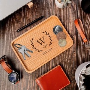 Puede incluir: Una bandeja de madera con una inscripción personalizada que incluye la letra "W", el nombre "wilson" y la fecha "EST. 1991". La bandeja está decorada con un diseño de corona de laurel y tiene un acabado de madera clara. La bandeja está llena de llaves y monedas.