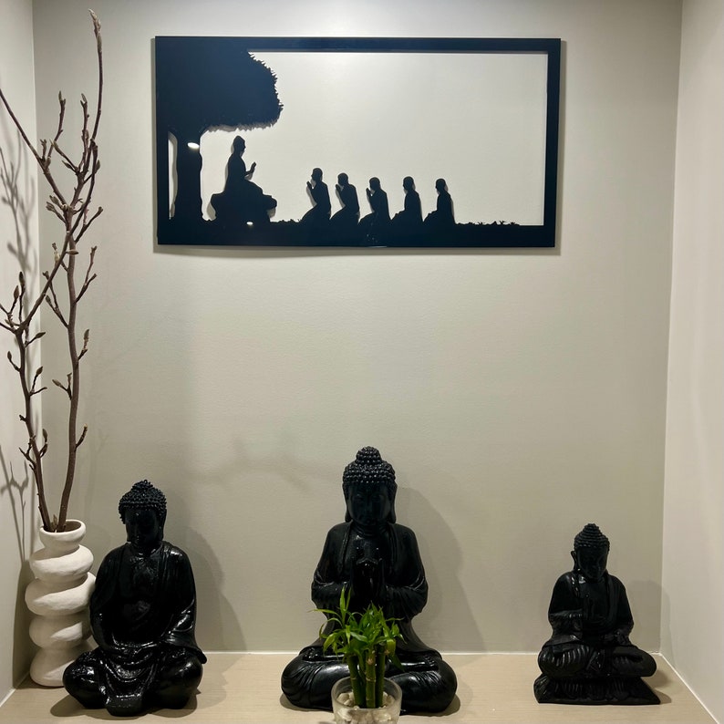 May include: Black and white silhouette wall art depicting a group of people meditating. Three black Buddha statues are placed in front of the art, with a small potted plant in the centre.