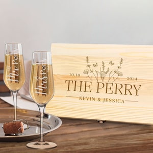 Könnte beinhalten: Zwei Champagnergläser mit den eingravierten Namen "Mr Perry" und "Mrs Perry", gefüllt mit einem prickelnden Getränk. Eine Holzkiste mit "The Perry" und Hochzeitsdatum im Hintergrund. Ein Korken und ein Flaschenöffner liegen auf einem silbernen Tablett.