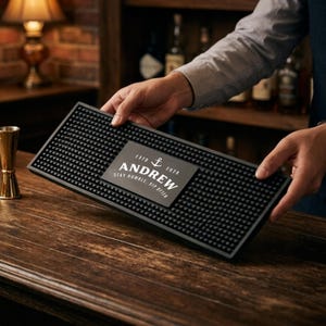 Peut inclure: Un tapis de bar rectangulaire en caoutchouc noir avec une surface texturée de petits picots en relief. Au centre, une plaque rectangulaire gris foncé présente une icône d'ancre, le texte ESTO 2026, le nom ANDREW et STAY HUMBLE, SIP OFTEN en blanc.