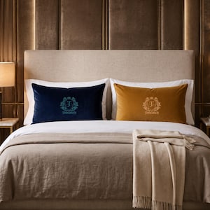May include: Two rectangular velvet throw pillows placed on a bed. One pillow is dark blue with a light blue embroidered crest featuring the letter T and the text TIMMERMAN. The other pillow is mustard yellow with a gold embroidered crest.