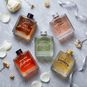 May include: Five square glass bottles filled with coloured liquids, each featuring custom printed labels. The bottles contain various beverages labelled Dry Martini, Piña Colada, Limoncello Mocktail, Mr & Mrs Anderson, and Andrew, with dates and text.