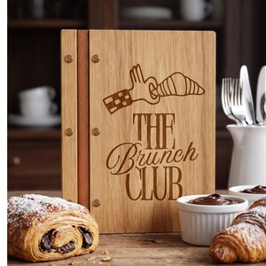 Puede incluir: Un portamenús rectangular de madera con un lomo de cuero marrón sujeto por tres remaches metálicos. El panel frontal está grabado con láser con un gráfico de una mano sosteniendo un cruasán y el texto "THE Brunch CLUB" en una fuente estilizada.