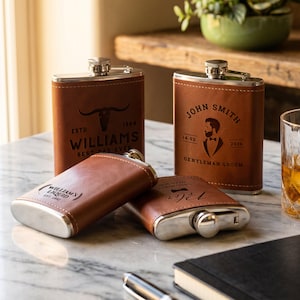 May include: Four brown leather-wrapped metal flasks with silver caps. Two stand upright, one featuring a bull skull graphic with the text "WILLIAMS ESTD 1984 BEST DAD EVER," and the other showing a groom silhouette with "JOHN SMITH 14.02 2026 GENTLEMAN GROOM."