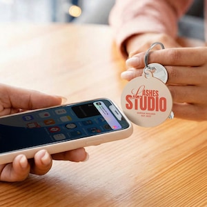 May include: A round keychain with the text "Lashes Studio" in red script and block letters, with "Sophia Walker Est. 2027" below. The keychain is held near a smartphone in a light-colored case.