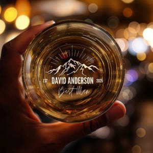 May include: A clear glass with a mountain range design and the text "EST DAVID ANDERSON 2025 Best Man" etched into the glass.