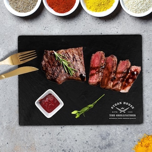 Puede incluir: Una tabla de servir de pizarra negra con un gran bistec y varios bisteces más pequeños, un pequeño tazón blanco con salsa roja y una ramita de romero. La tabla tiene el texto "Steak House" y "The Grillfather" impreso en ella.