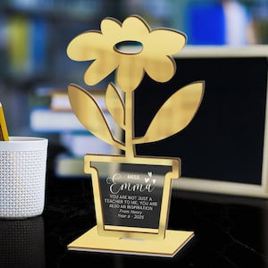 May include: A gold-coloured flower-shaped desk ornament. The flower has a cutout design and is mounted on a rectangular base. The flower pot section has the name "Emma" and a message: "YOU ARE NOT JUST A TEACHER TO ME, YOU ARE ALSO AN INSPIRATION From Henry Year 6 - 2025."