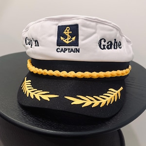 May include: A white and black captain's hat with gold accents. The hat features the name "Cap'n Gabe" embroidered in black, along with an anchor emblem and the word "CAPTAIN". A gold rope trim and gold leaf design complete the nautical theme.