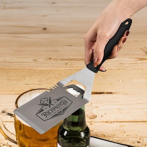 Puede incluir: Una espátula de parrilla de plata con un abridor de botellas integrado y un borde dentado. La espátula tiene un mango negro y está grabada con el texto "EST. 1994 RICHMOND GRILL MASTERS & CO."