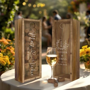 May include: Two wooden wine bottle boxes with glass inserts. The box on the left has a white inscription that says "Matt & Vickie 16.10.2030" and is filled with wine corks. The box on the right has a white inscription that says "E&K Eleanor & Korina 02.05.2025" with a floral design. A glass of white wine sits in front of the boxes.