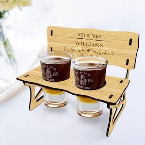 May include: A wooden bench shot glass holder with two shot glasses. The bench has a personalized inscription that reads "MR & MRS WILLIAMS 09.12.2025". The shot glasses are engraved with "Groom to be 2025 Willow" and "Bride to be 2025 Victoria".