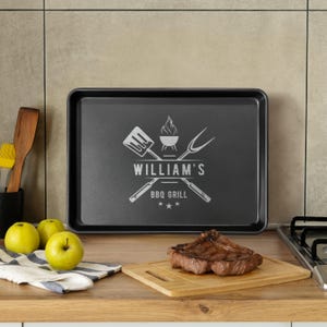 以下が含まれることがあります： 「WILLIAM'S BBQ GRILL」の文字と焼き網のグラフィックが入った黒い長方形のBBQグリル用トレイ。調理済みのステーキが木製のまな板の上に置かれています。青リンゴとストライプの布も見えます。