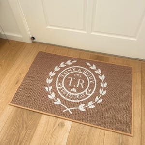 May include: A brown doormat with a white wreath design featuring the text "Tony & Ruby" and "T.R. EstD 2023".