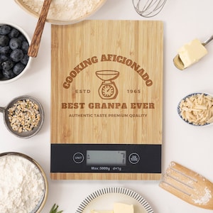 Może przedstawiać: Brązowa drewniana waga kuchenna z czarnym wyświetlaczem cyfrowym. Waga ma grawerowane laserowo wzory z napisem "Cooking Aficionado Est. 1965 Best Granpa Ever Authentic Taste Premium Quality".