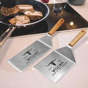 Może przedstawiać: Dwie srebrne metalowe szpatułki z drewnianymi uchwytami. Szpatułki mają czarny wzór głowy byka i tekst "ESTD 1984 WILLIAM CHIEF OF THE CHARCOAL".