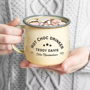 Puede incluir: Una taza de esmalte color crema llena de chocolate caliente y malvaviscos. La taza tiene un borde gris y un asa verde. El texto en la taza dice "HOT CHOC DRINKER" con un nombre y "Extra Marshmallows" debajo. El año 2024 también está impreso en la taza.