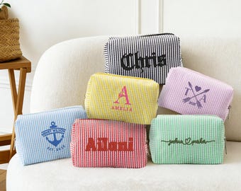 Puede incluir: Una colección de neceseres personalizados en varios colores, incluyendo azul, amarillo, rojo, morado y verde. Cada neceser presenta un nombre o monograma único en un color contrastante, con una textura a rayas. Los neceseres están colocados sobre un sofá blanco.