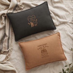 May include: A rectangular charcoal gray pillow with a gold heart-shaped graphic featuring the text Jason & Ema 11.08.2026 and a tan pillow with a brown graphic of a house and the text CONGRATULATIONS Jason & Ema EST. 2026 New Home New Memories.