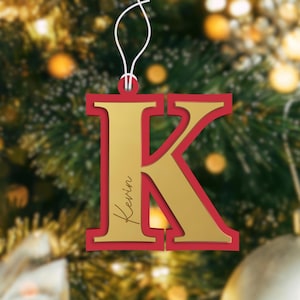 Puede incluir: Adorno navideño de la letra K dorada con borde rojo. El adorno tiene el nombre "Kevin" inscrito en él.