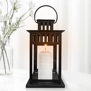 May include: Black metal lantern with a glass panel and a white candle inside. The candle has an inscription that reads "In loving memory of Karen Margare 1996-2022 He will be remembered by his wife and daughter to many others family and colleagues. Monday, March 28th."