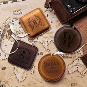 May include: Several personalised leather tape measures in different shapes and shades of brown. The tape measures are engraved with names and initials. One is square and tan, another is round and dark brown, and a third is square and dark brown.