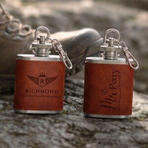 May include: Two stainless steel pocket flasks wrapped in brown textured leather. Each flask features a silver screw-top cap with a metal keychain attachment. One flask is engraved with a winged crest and the text RICHMOND A GENTLEMAN'S ESSENTIALS, while the other displays the text Mr Perry and the date 10.10.2026.
