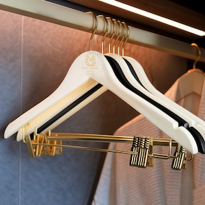 Puede incluir: Un conjunto de perchas de madera color crema con ganchos y pinzas doradas. Las perchas tienen una barra negra y el texto "HAPPY BIRTHDAY OLIVA" en dorado. Las perchas cuelgan de una barra dorada.