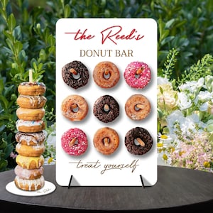 Pode incluir: Uma placa branca com o texto "the Reed's DONUT BAR treat yourself" em escrita marrom. A placa tem uma parede de donuts com nove mini donuts em várias cores e coberturas.