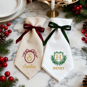 May include: Two personalised wine bottle bags. One is beige with a burgundy ribbon and the name "Sophia" embroidered in gold. The other is white with a green ribbon and the name "Henry" embroidered in gold. Both have decorative embroidered designs.