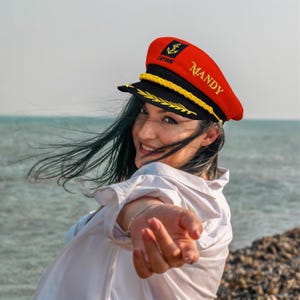 May include: A red and black captain's hat with gold trim and the name "Mandy" embroidered on the side. The hat features a gold anchor emblem and the word "CAPTAIN".