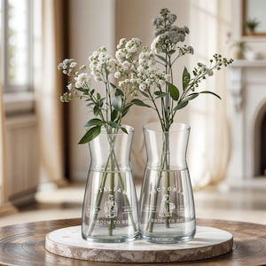 Puede incluir: Dos jarrones de vidrio transparente con flores blancas. Los jarrones están grabados con las palabras "Groom to Be" y "Bride to Be" y los nombres "William" y "Victoria".
