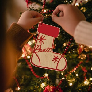 Op de afbeelding: Een houten kerstsok ornament met de naam "Brett" geschreven in rood cursief. De sok is versierd met rode sterren en heeft een rode vilt top.