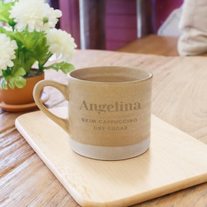 May include: A beige ceramic mug with a handle, personalized with the name "Angelina" and coffee order details. The mug sits on a wooden tray, with a white base and is near a potted plant.