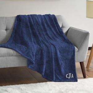 Può includere: Una coperta in pile blu navy con le iniziali "CJA" ricamate in bianco nell'angolo in basso a destra.
