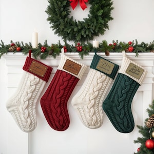 Pode incluir: Quatro meias de Natal personalizadas com um design de tricô trançado. As meias estão penduradas em uma lareira branca. As meias são brancas, vermelhas, verdes e creme com etiquetas de couro douradas. Os nomes nas etiquetas são "Sophie's", "Belinda", "Mrs. Harrington" e "Sacha Dubois".