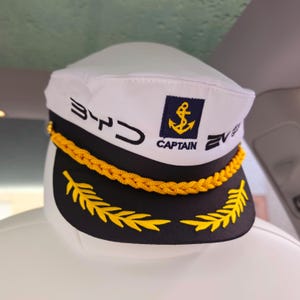 May include: White captain's hat with a black brim and gold accents. The hat features the word "BYD" in black, a navy blue patch with a gold anchor, and the word "CAPTAIN". A gold rope trim and gold leaf embroidery complete the design.