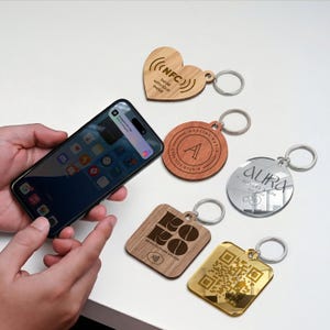May include: A collection of keychains in various shapes and materials, including wood and mirrored metal. One keychain is heart-shaped with "NFC" and "Tap me with your phone" text. Other keychains feature logos and a QR code. A smartphone is also in the image.