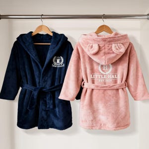 May include: Two plush hooded bathrobes hanging on wooden hangers. One is navy blue with a white emblem that reads "Henry's Fine Adventures". The other is pink with bear ears on the hood and a white emblem that reads "Little Hall Est. 2025".