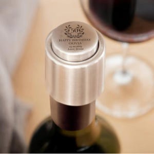 May include: A silver wine bottle stopper with the text "HAPPY BIRTHDAY OLIVIA 25.10.2025 Love, Jessie" and a decorative wreath with the number 50. The stopper is on a wine bottle, with a glass of red wine in the background.
